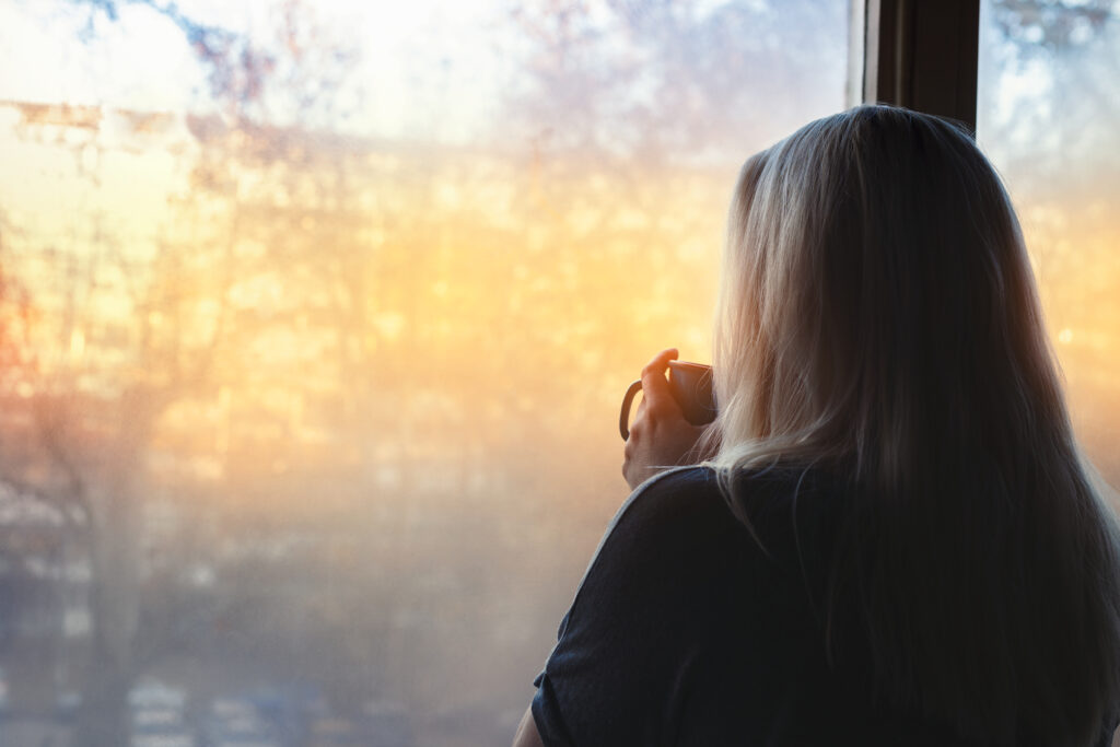 woman standing by window, coffee cup in hands, looking out into morning light