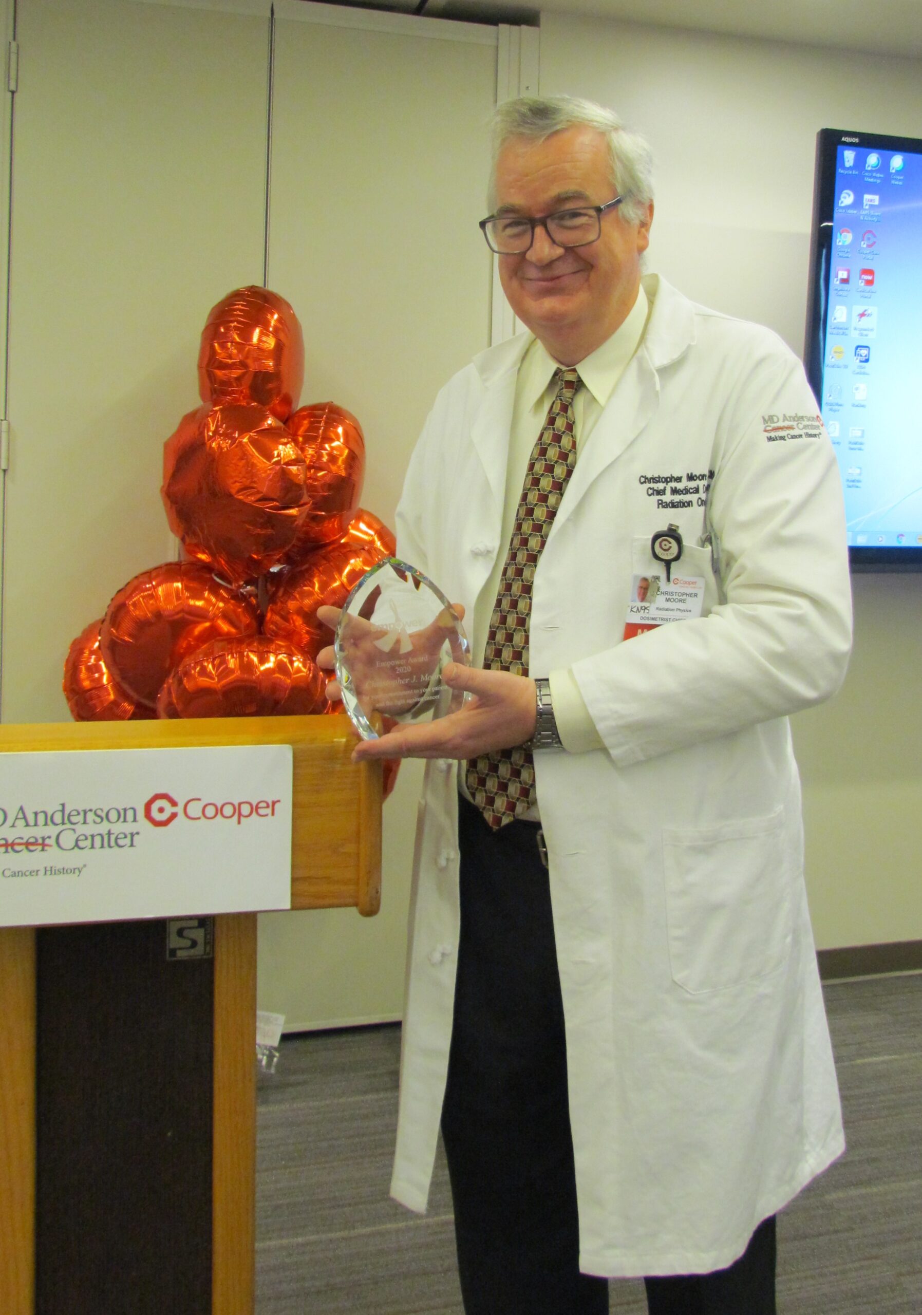 Christopher J. Moore, BS, CMD, a Medical Dosimetrist at MD Anderson Cancer Center at Cooper, holds the 2020 Radiation Therapy Power Award next to an MD Anderson Cancer Center at Cooper lectern.