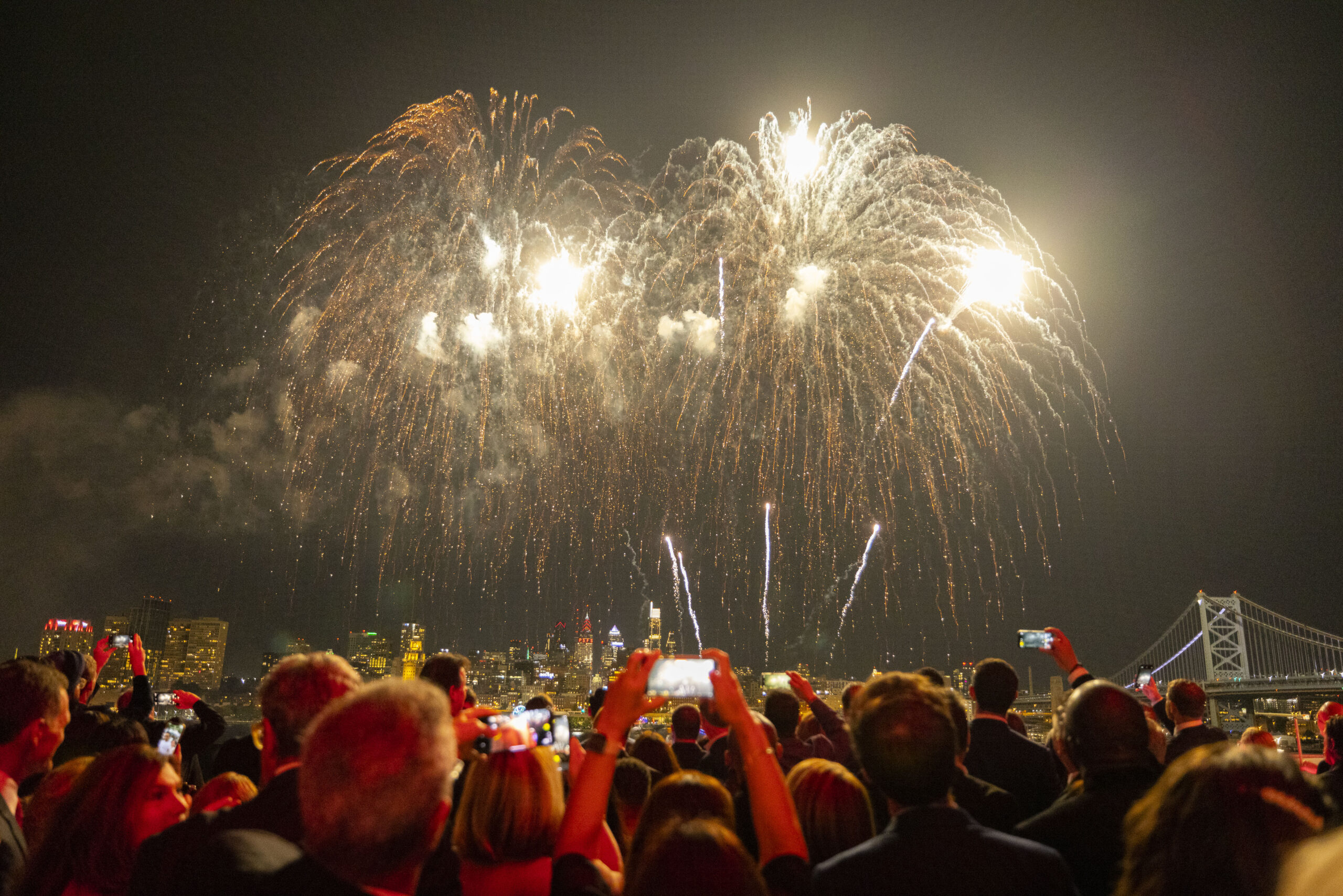 Fireworks explode in the night sky above a large crowd with the Ben Franklin Bridge in the distance.