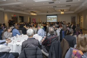 Wide angle shot from the back of a crowded room of individuals seated at tables.